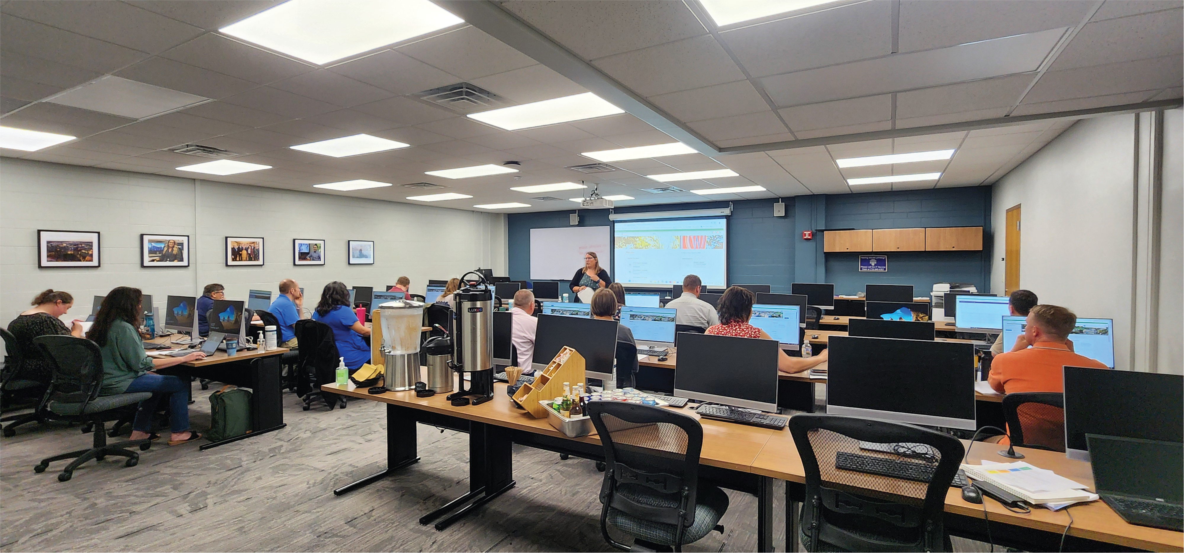 Staff sit behind rows of computers in a conference room with a large projection screen at the front.