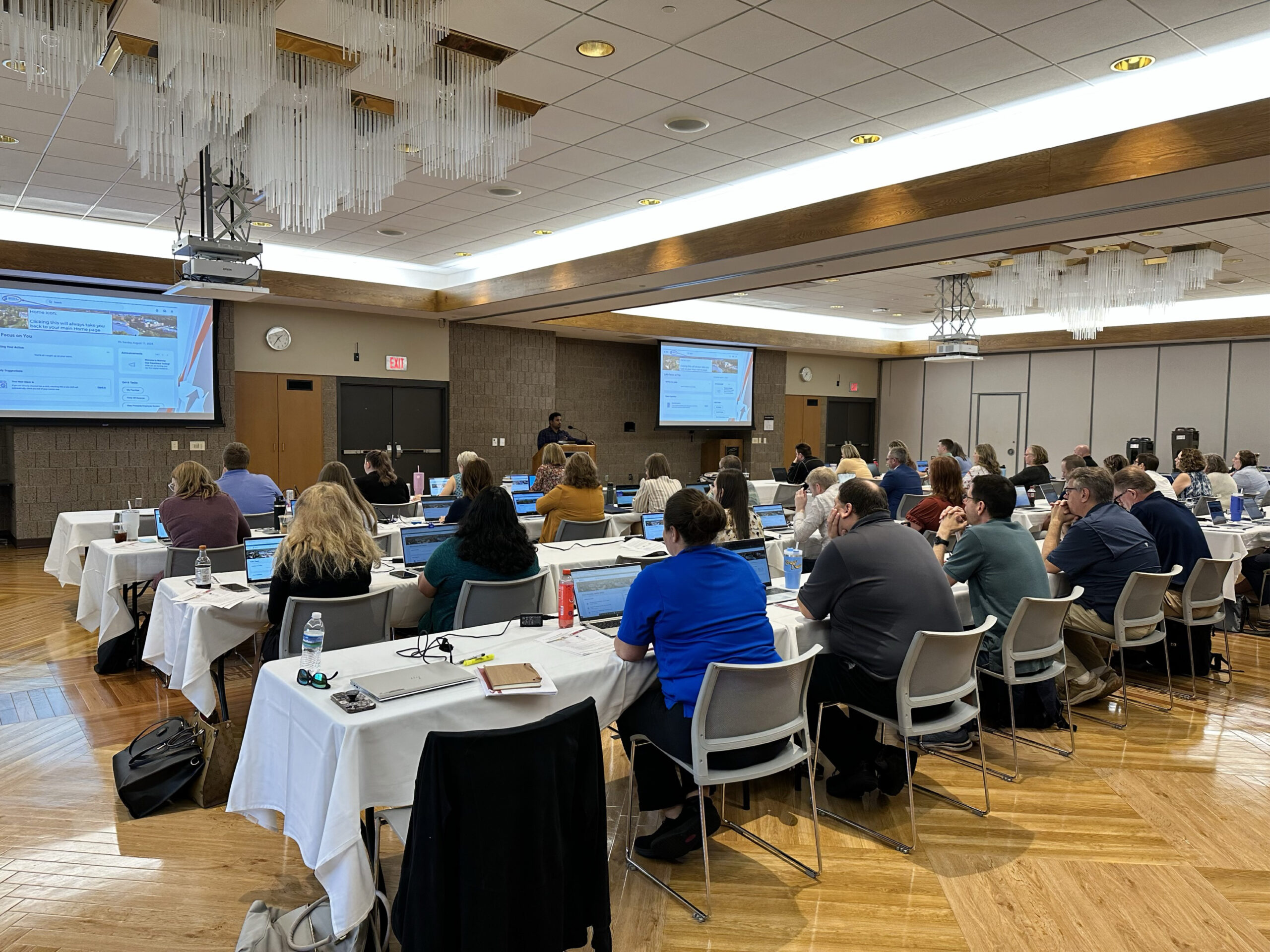 Dozens of staff sit behind computers in a large conference room with two projection screens at the front.