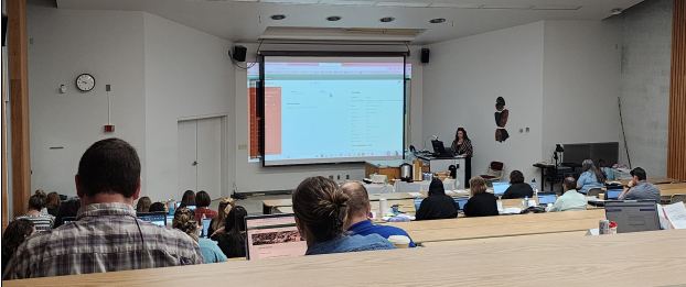 A conference room is filled with UW staff members who face a large projection screen with a presenter at the front of the room.